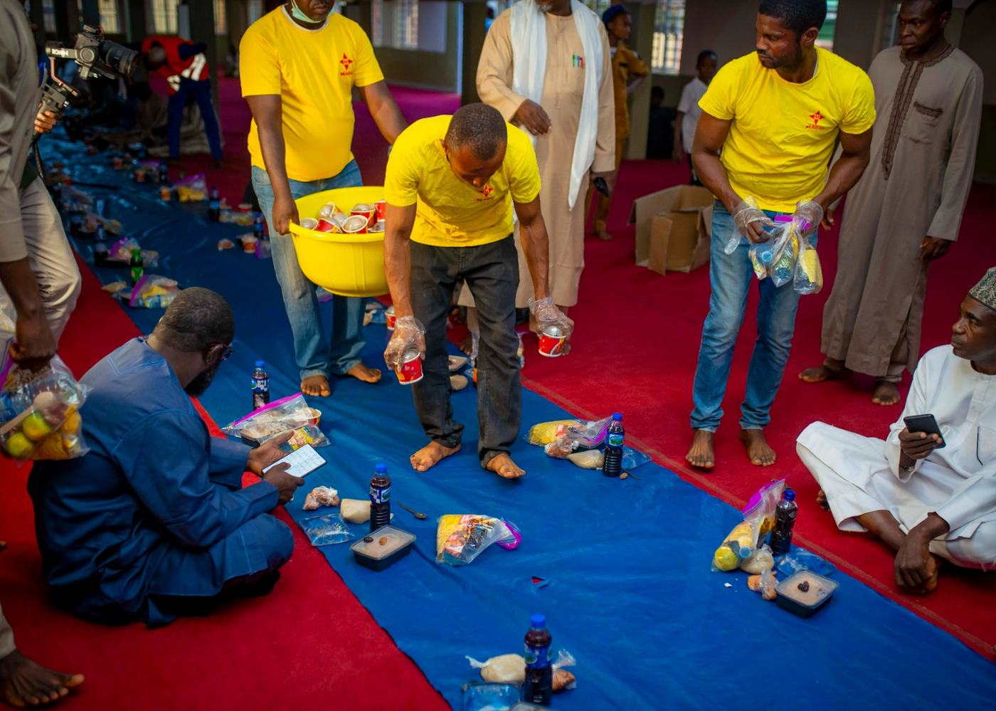 Team members distributing packs to guests seated on mats inside a community hall.