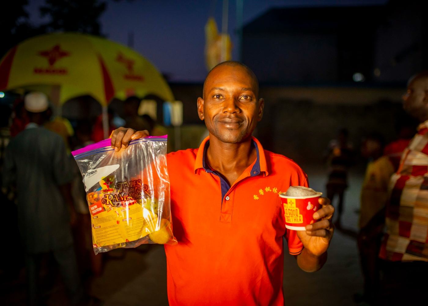 A guest in an orange shirt holding a Tales of Ramadan pack and a branded cup.