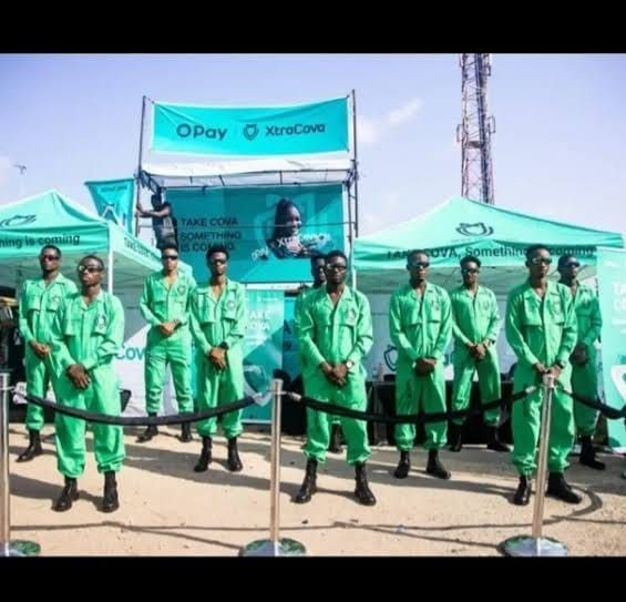 Brand ambassadors in green coveralls and caps standing behind a rope barrier.